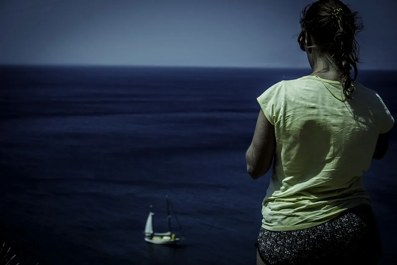 Frau blickt aufs Meer, ein Segelboot am Horizont
